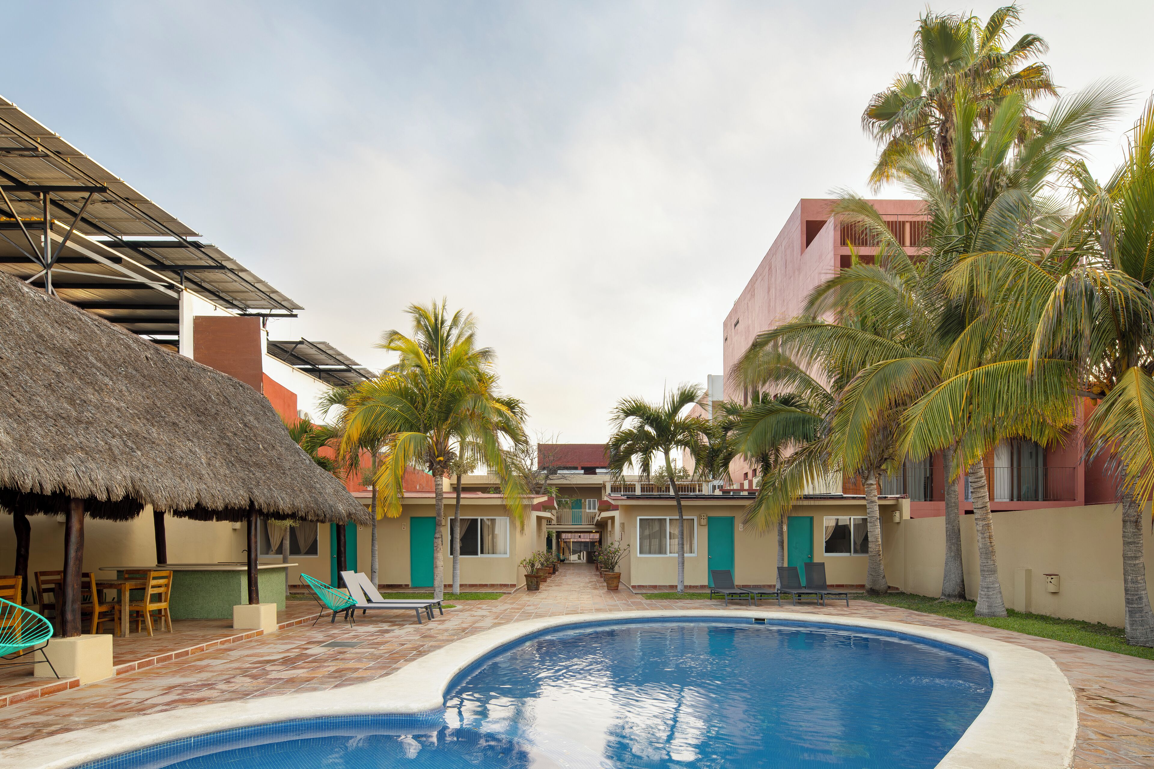 Una piscina con sillas de descanso y una estructura de techo de paja, rodeada de palmeras y edificios con puertas verdes.