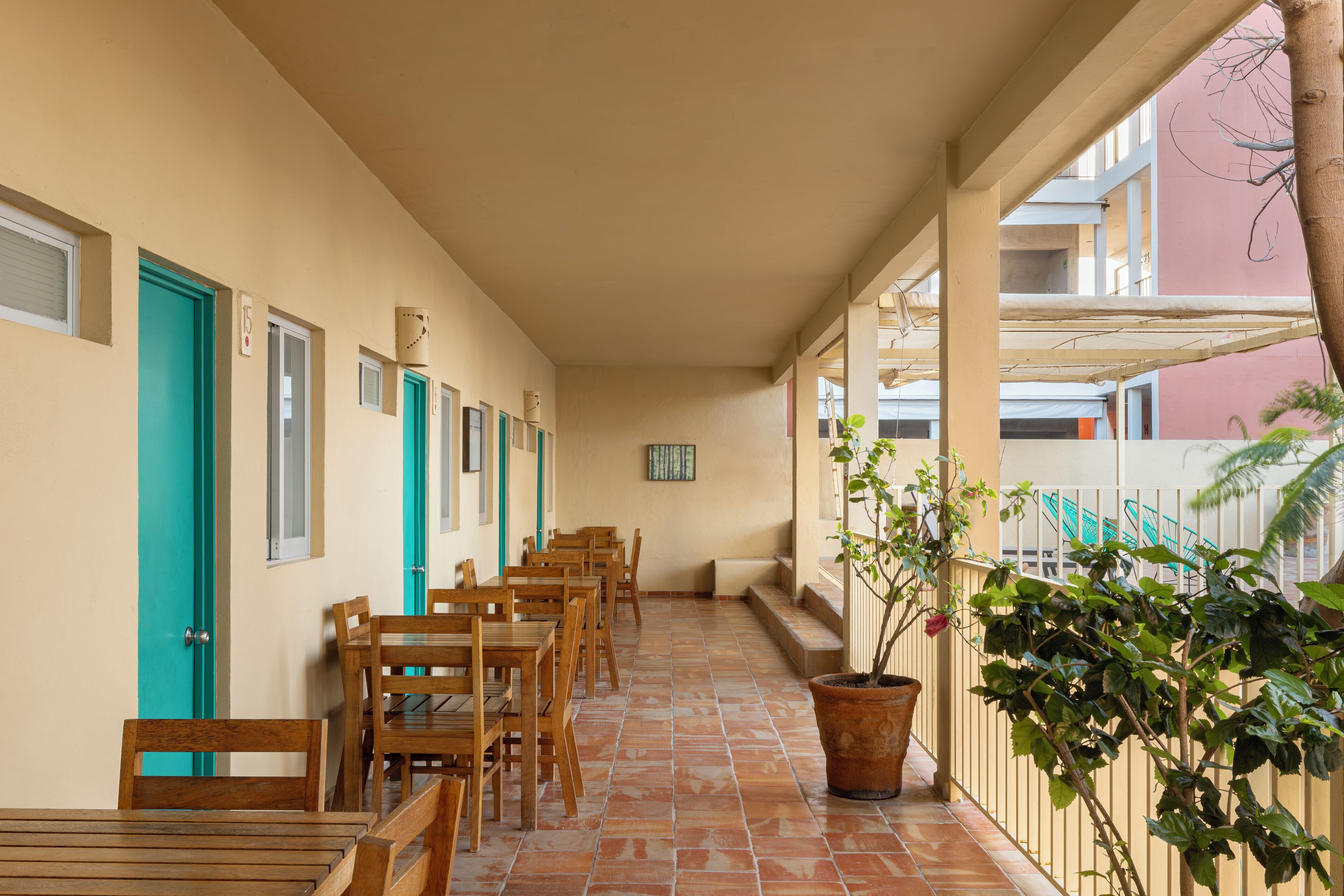 A corridor with wooden tables and chairs, numbered doors, and a potted plant.