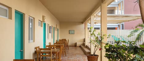 A corridor with wooden tables and chairs, numbered doors, and a potted plant.
