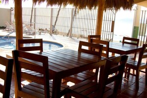 Uma área na piscina com mesas e cadeiras de madeira, um telhado de palha e vista para a piscina e a praia.