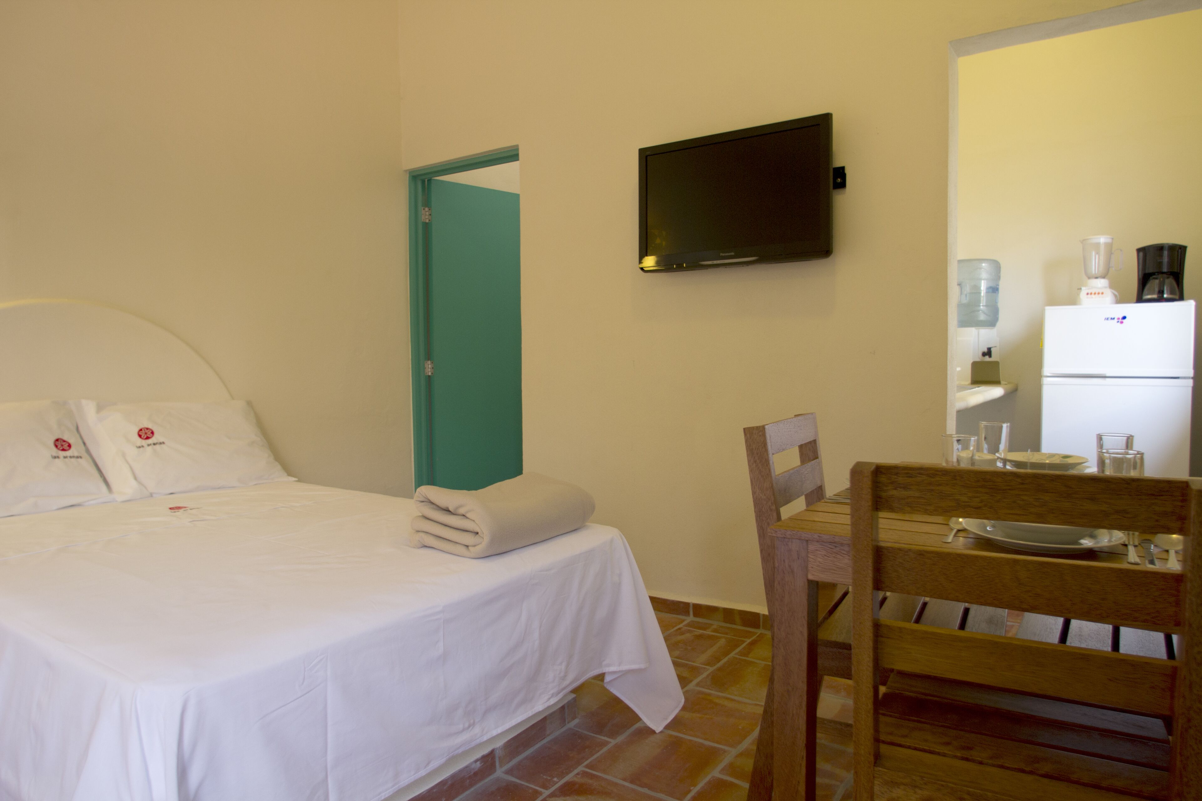 Habitación de hotel con cama, televisor en la pared, una mesita con una silla y un refrigerador.