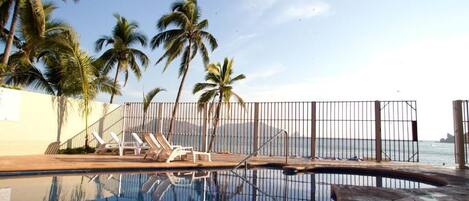 Piscina con sillas de descanso y palmeras, rodeada por una cerca.