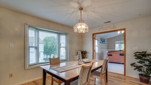 Un coin repas avec une table en bois, des chaises, un luminaire suspendu et une vue sur la cuisine à travers une porte ouverte.