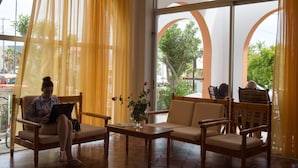 A room with a large window, a chair, a sofa, and a table with a laptop and a vase of flowers. - Laura (Kos)