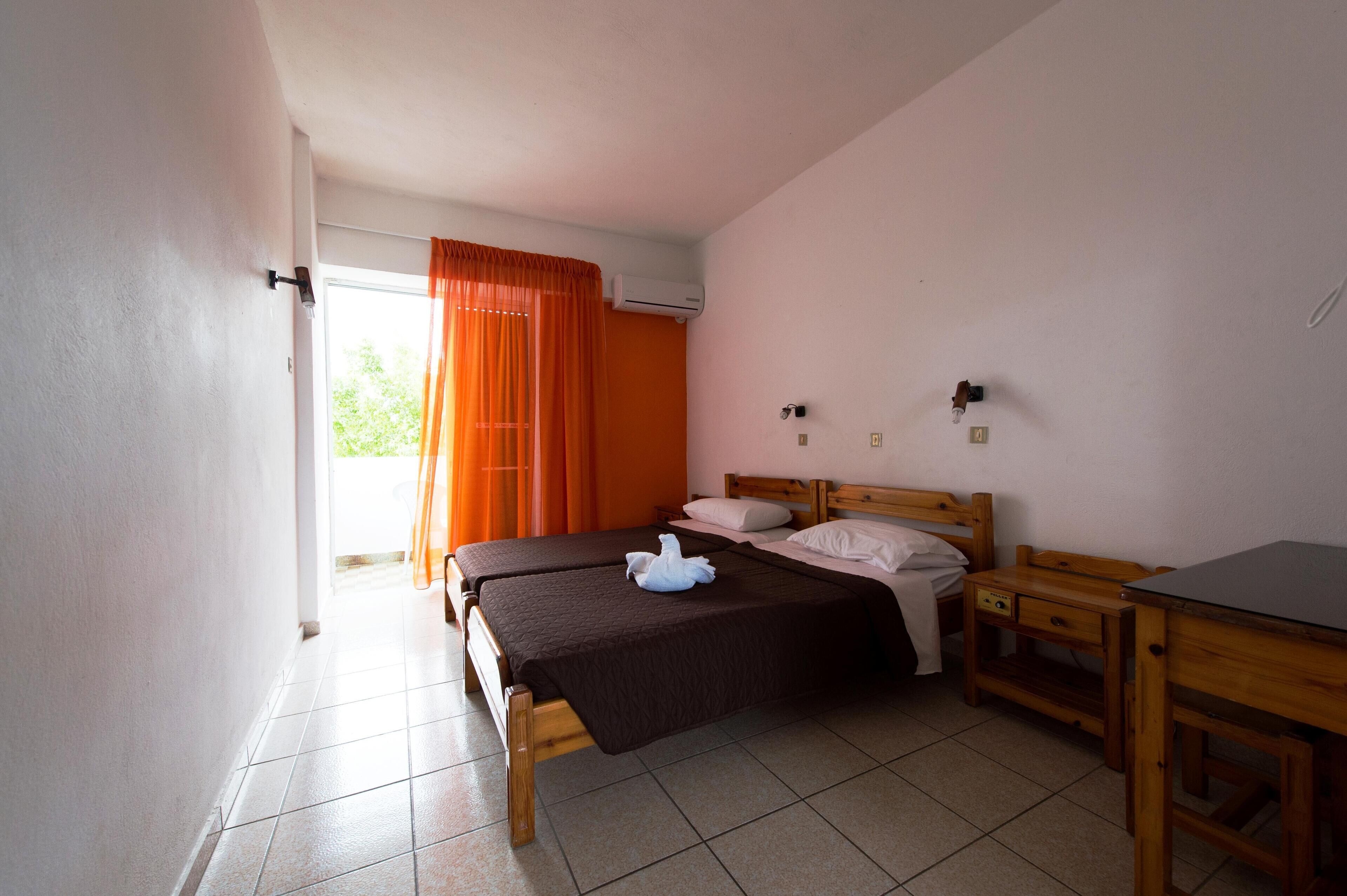 Quarto com duas camas, ar condicionado, escrivaninha de madeira e janela com cortinas laranja.