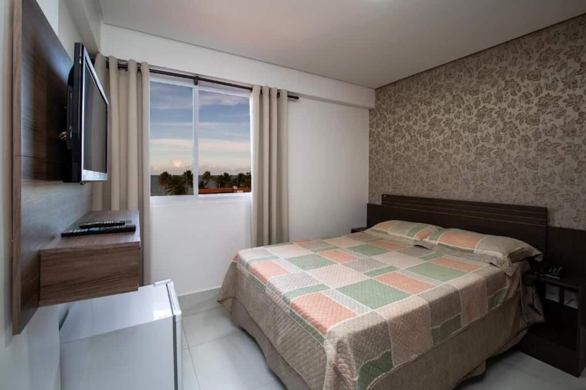 A bedroom with a bed, a TV, a window with curtains, and a patterned wall.