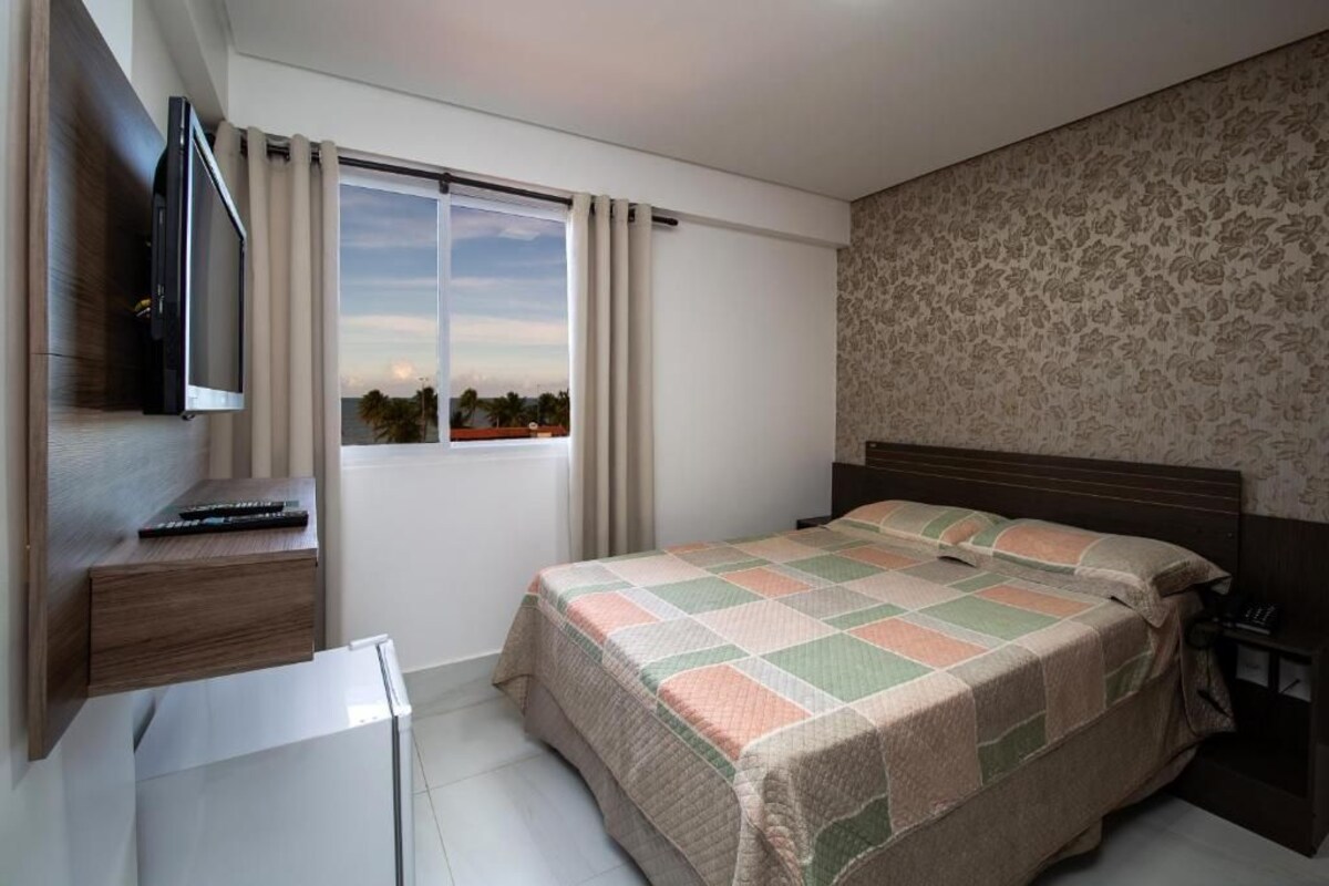 Une chambre à coucher avec un lit, une télévision, une fenêtre avec des rideaux et un mur décoré d’un motif.
