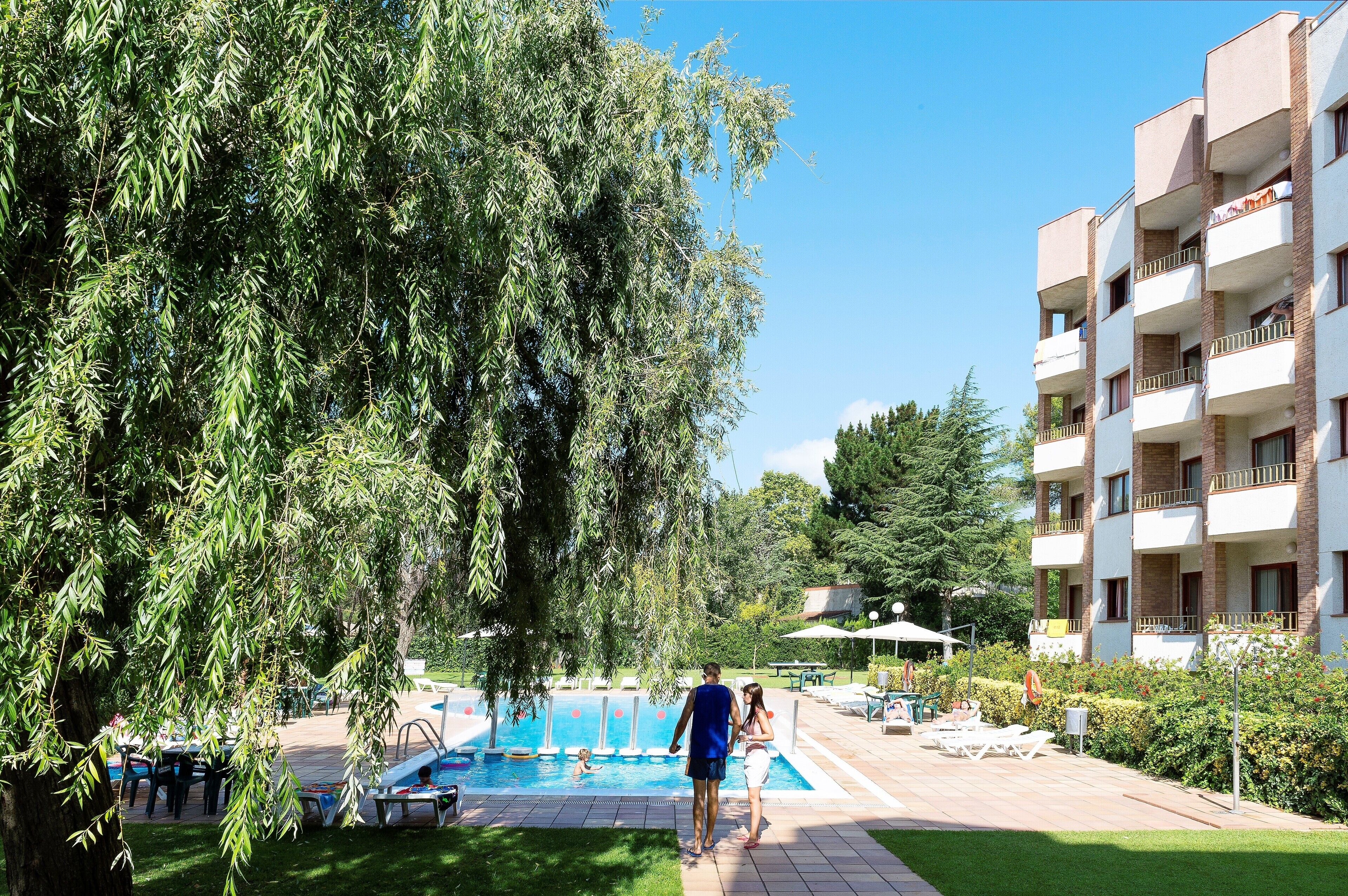 A swimming pool with lounge chairs, a building with balconies, and a large tree.