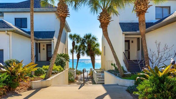 A beachfront property with a concrete pathway leading to a gated entrance, surrounded by palm trees and various shrubs. - Swell Life - Private Beach- 4 Bikes (Santa Rosa Beach)