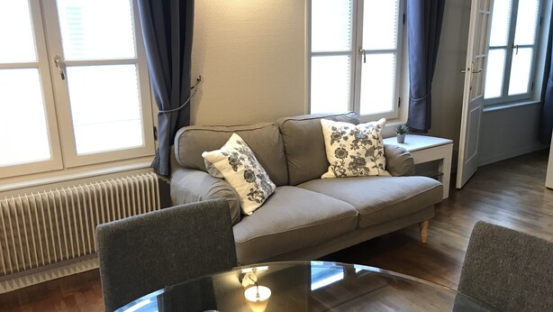 Een woonkamer met een beige bank, twee bloemenkussens, een glazen salontafel en een klein bijzettafeltje.
