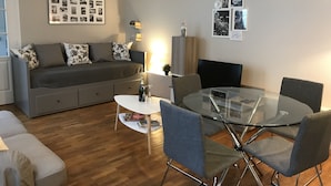 Een moderne woonkamer met een grijze bank, een glazen eettafel en enkele stoelen. Er is een boekenkast en een wandcollage met foto's.