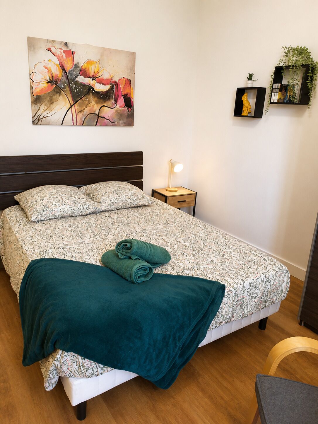 A neatly made bed with patterned bedding, a green throw blanket, a bedside lamp, a small plant, and a framed picture on the wall.