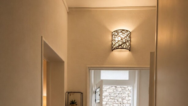 Un couloir avec un mur en pierre, une porte donnant sur l’extérieur, un paillasson noir à l’entrée et un luminaire fixé au mur.