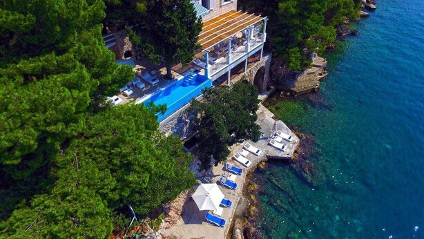 Eine Luftaufnahme eines Anwesens an der Küste mit Swimmingpool, überdachter Terrasse und Liegestühlen entlang des Ufers.