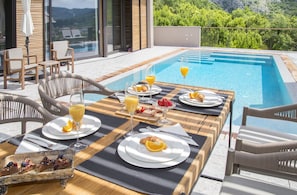 Une table à manger avec des aliments pour le petit-déjeuner, une piscine et des sièges d’extérieur.