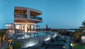 A modern multi-story house with a swimming pool, outdoor seating, and a view of the sea.