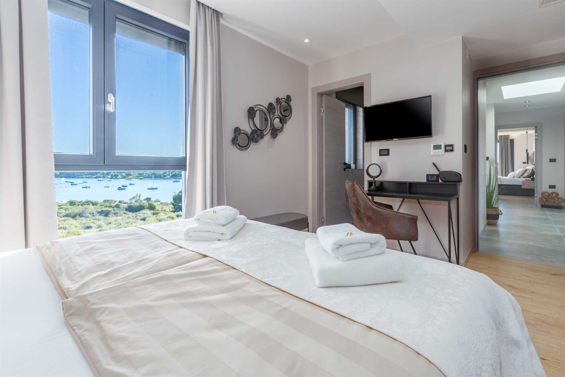 A modern hotel room with a large bed, a desk, a TV, and a view of the sea.