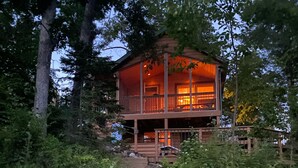 A wooden cabin with a balcony, surrounded by dense foliage and a wooden staircase leading up to it. - Secluded Waterfront Cabin on hiking trails! Lake on one side river on the other. (Luck)