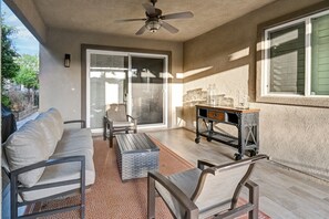 A covered patio area with a ceiling fan, a sliding glass door leading outside, a cushioned outdoor sofa, a small table with a glass jar, and a metal cart.