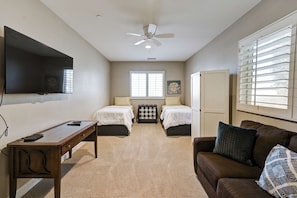 A room with two beds, a desk, a television, a ceiling fan, and a window with blinds.