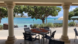 Ein überdachter Essbereich im Freien mit Blick auf das Meer, ausgestattet mit einem Tisch, Stühlen, einer Pergola und einem großen Baum.