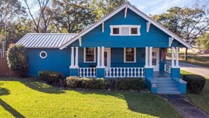 A single-story house with a gabled roof, a front porch with white railings, and a small front yard with grass and shrubs. - Fenced-In Yard & Nice Patio! Capital State Heights (Montgomery)