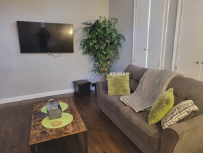 Una sala de estar con televisor de pantalla plana, una mesita de centro de madera, un sofá marrón con cojines, una planta en maceta y un altavoz negro pequeño.