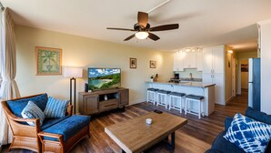 A modern living room with a sofa, coffee table, and a TV. There is a kitchen area with bar stools and a refrigerator.