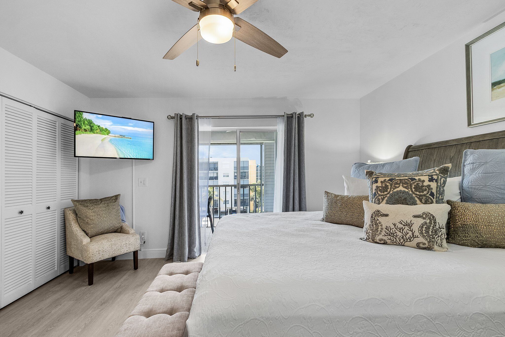 A bedroom with a bed, a chair, a ceiling fan, a TV, and a balcony with curtains.