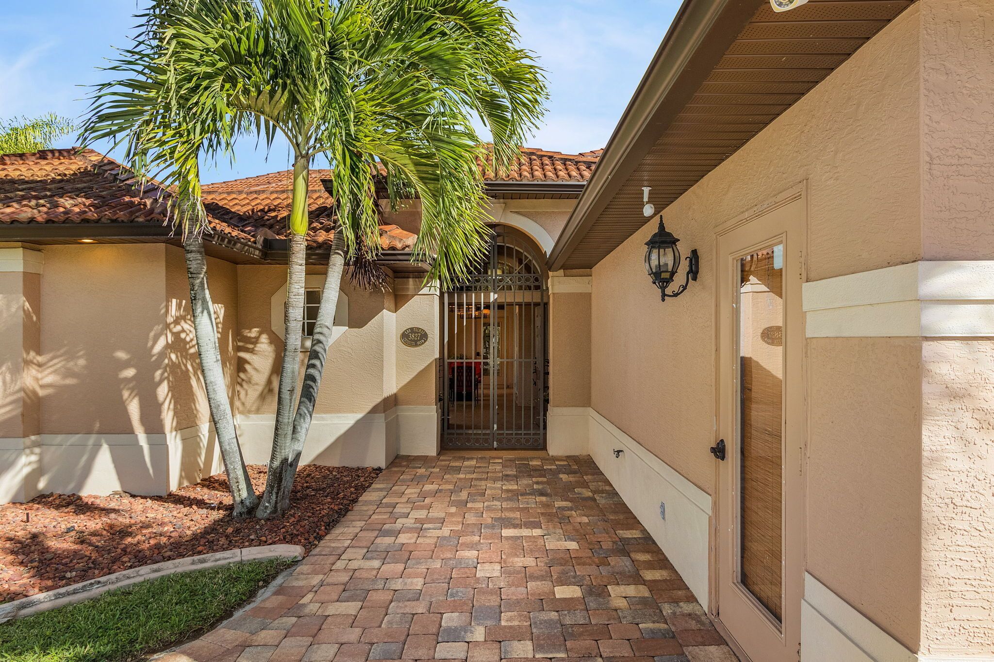 A covered walkway with a tiled floor, palm trees, and a gated entrance to a building with a beige facade and a tiled roof.