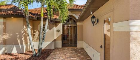 A covered walkway with a tiled floor, palm trees, and a gated entrance to a building with a beige facade and a tiled roof.