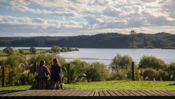 Dos personas sentadas en una cubierta de madera con vista a un lago y un paisaje boscoso al fondo.