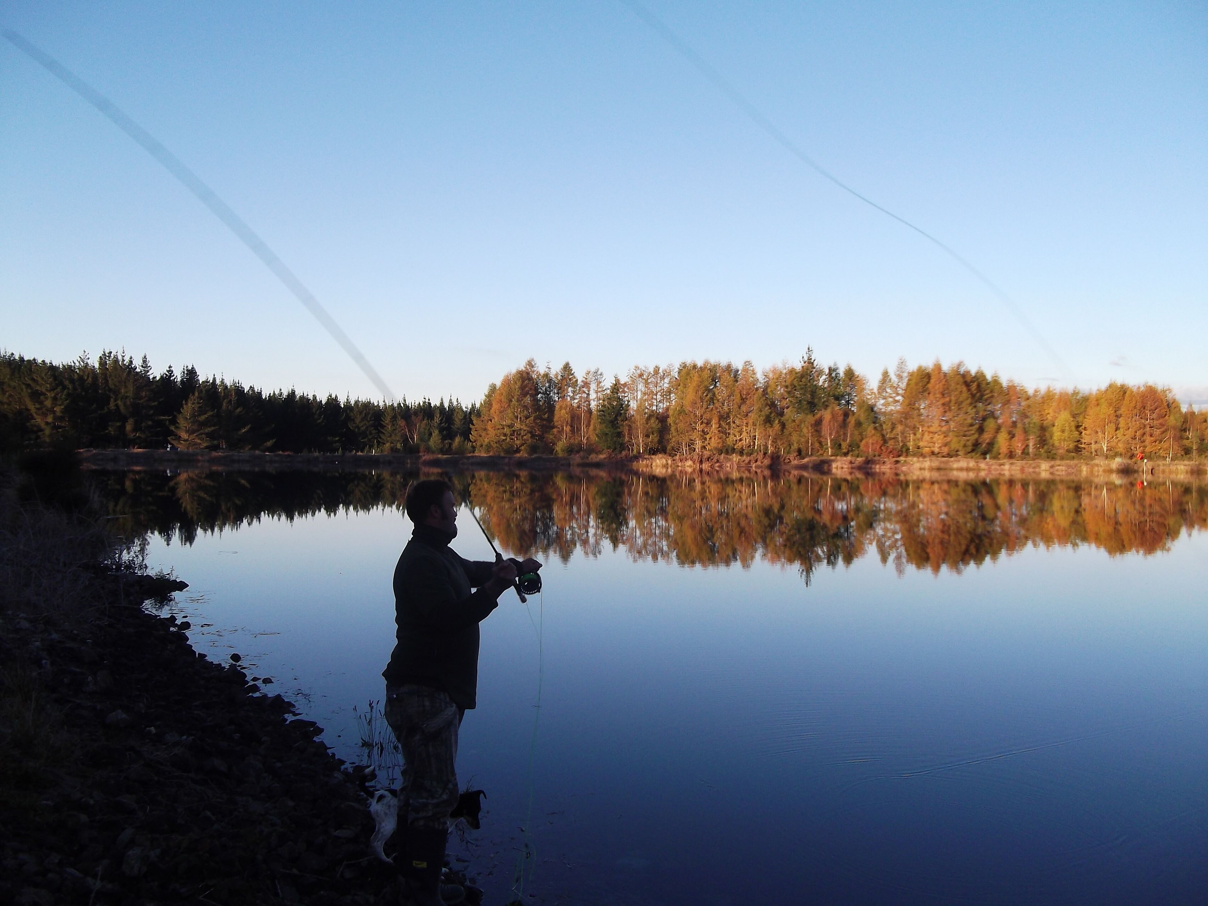 Eine Person beim Angeln an einem stillen See, mit Spiegelungen von Bäumen und blauem Himmel.