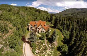 A large, two-story house with a red roof, surrounded by dense forest and hills.