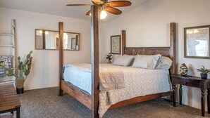 Une chambre avec un lit en bois, un ventilateur de plafond, une table de chevet, un cactus et un miroir au mur.