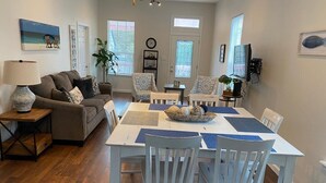 A dining area with a white table and chairs, a sofa, and a lamp. There are two windows with blinds, a ceiling fan, and a clock on the wall.