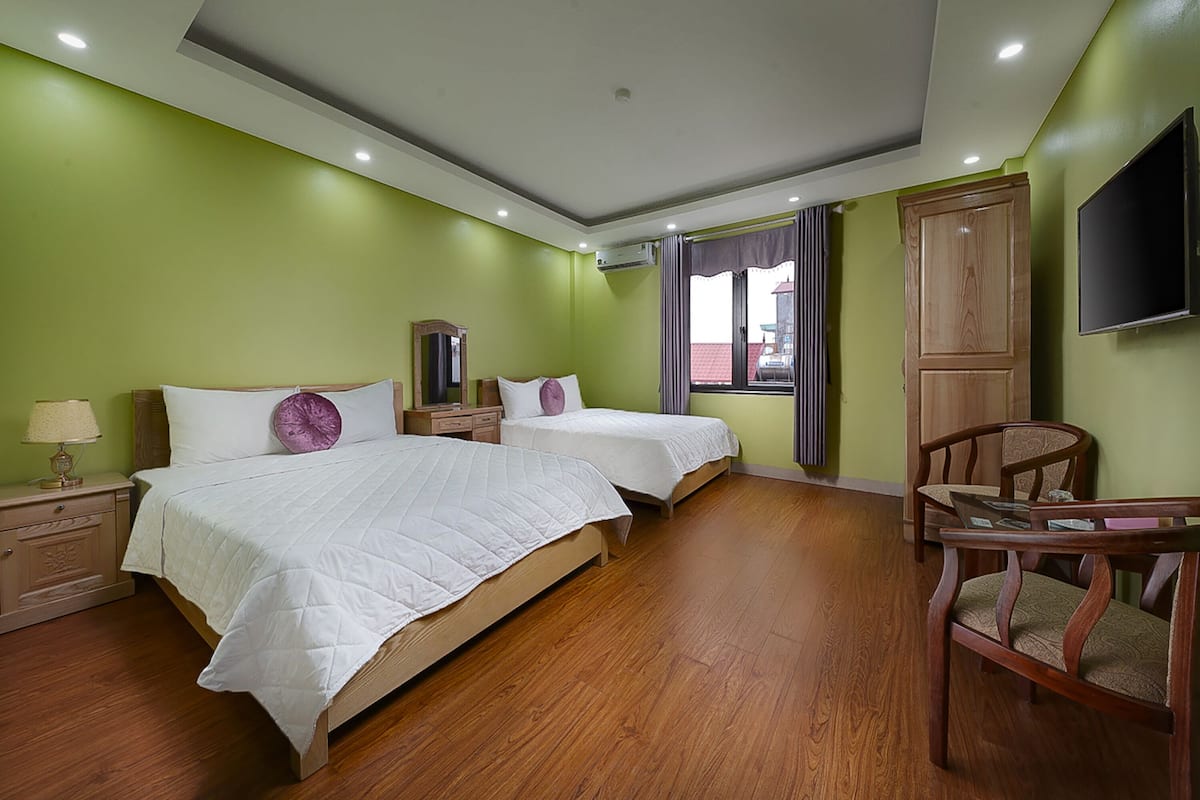 A hotel room with two beds, wooden flooring, a TV mounted on the wall, and a window with curtains.