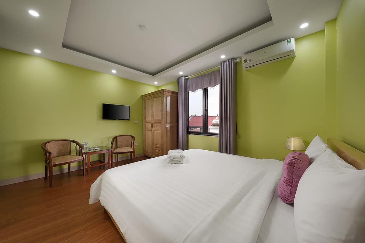 A hotel room with a bed, a TV, a desk with chairs, and an air conditioning unit.