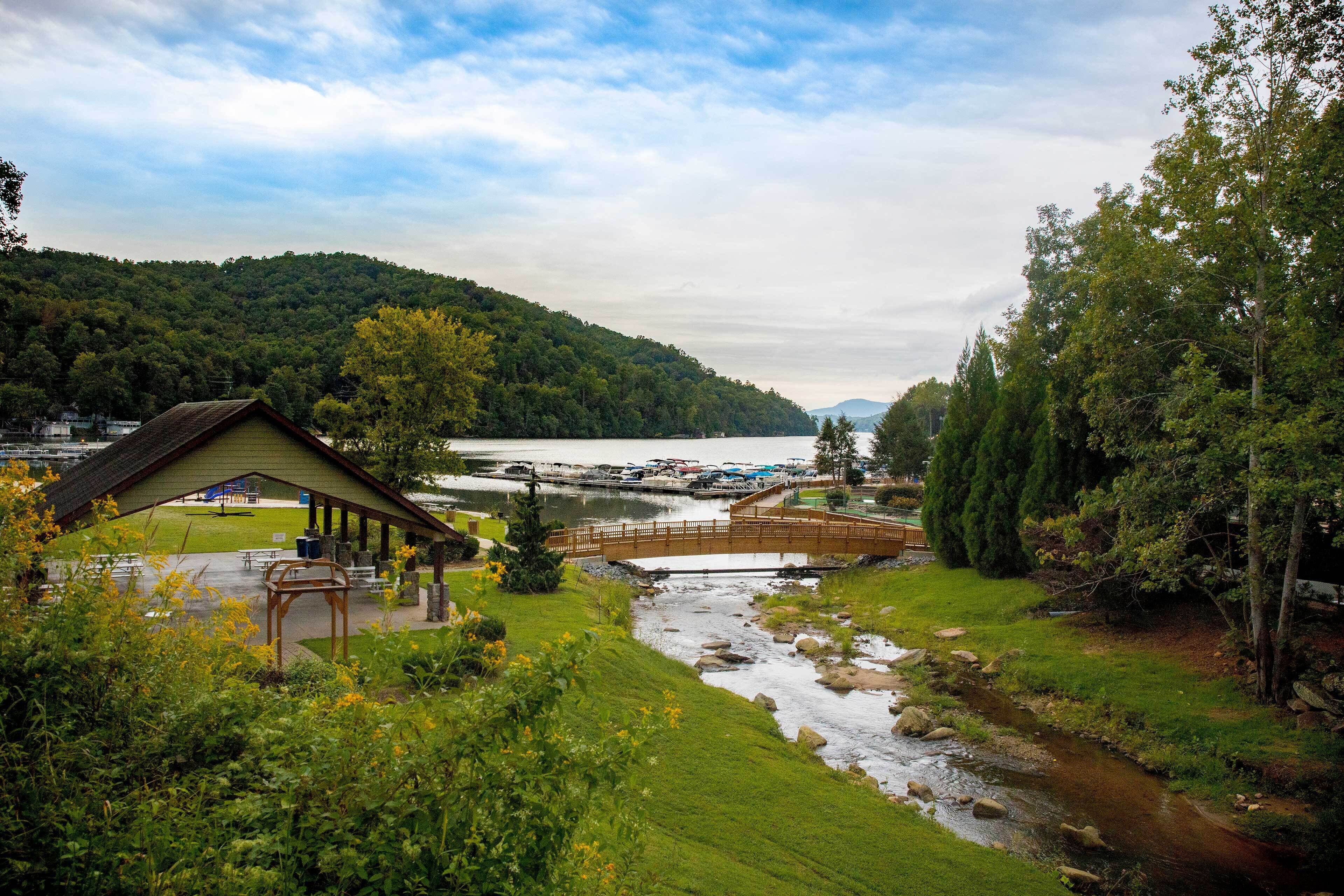 Un chalet au bord du lac, doté d’une terrasse couverte, d’un petit pont enjambant un ruisseau et offrant une vue sur les bateaux sur le lac.
