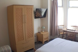 Habitación de hotel con un armario grande, un televisor montado en la pared, una mesita de noche con lámpara, una cama y un área de comedor con sillas y una mesa.