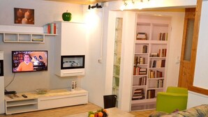 Ein Wohnzimmer mit Fernseher, Bücherregal, Couchtisch mit Obst und einem gemusterten Sofa.