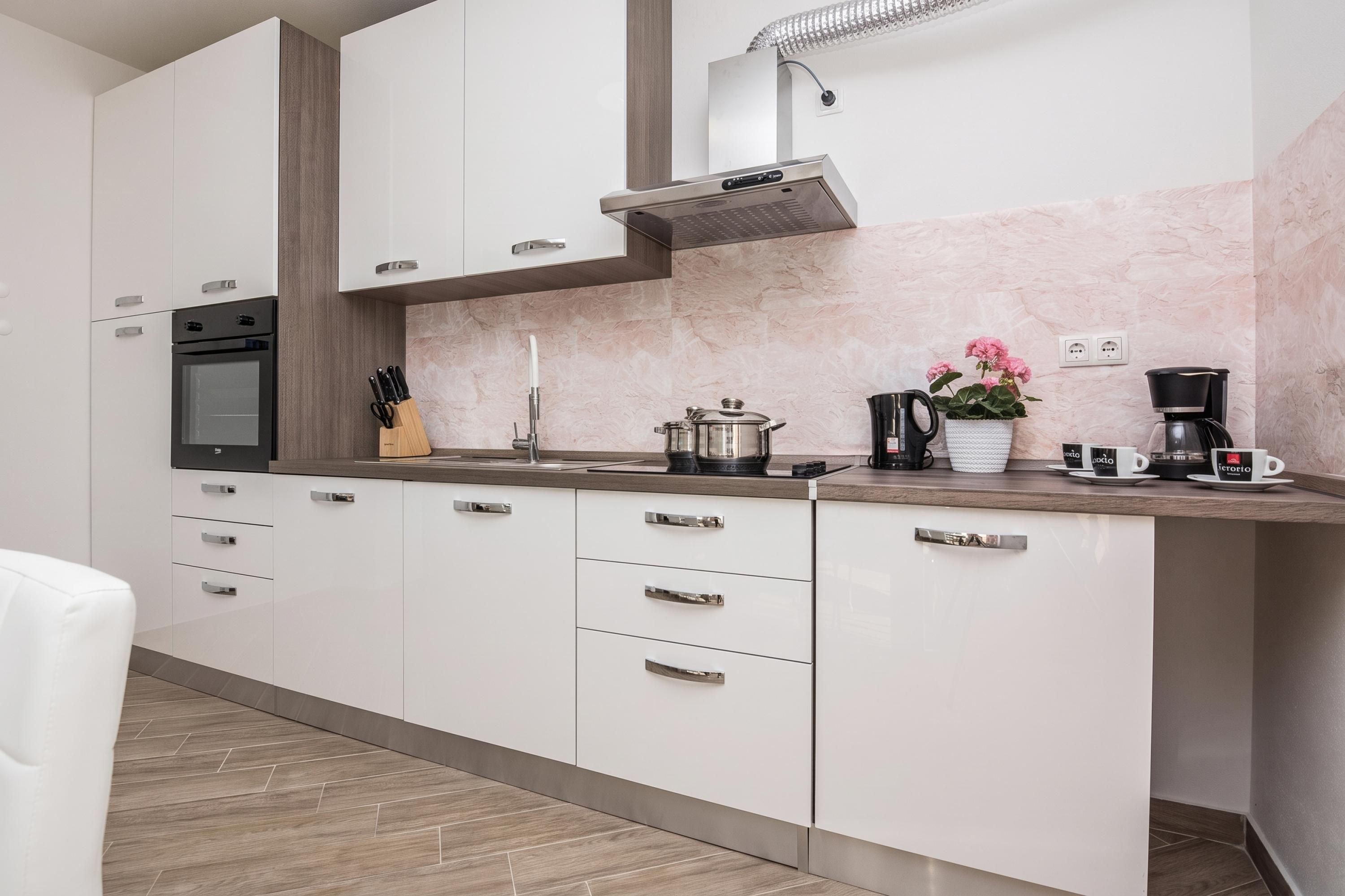 A modern kitchen with white cabinets, a built-in oven, a stainless steel range hood, and a marble backsplash.