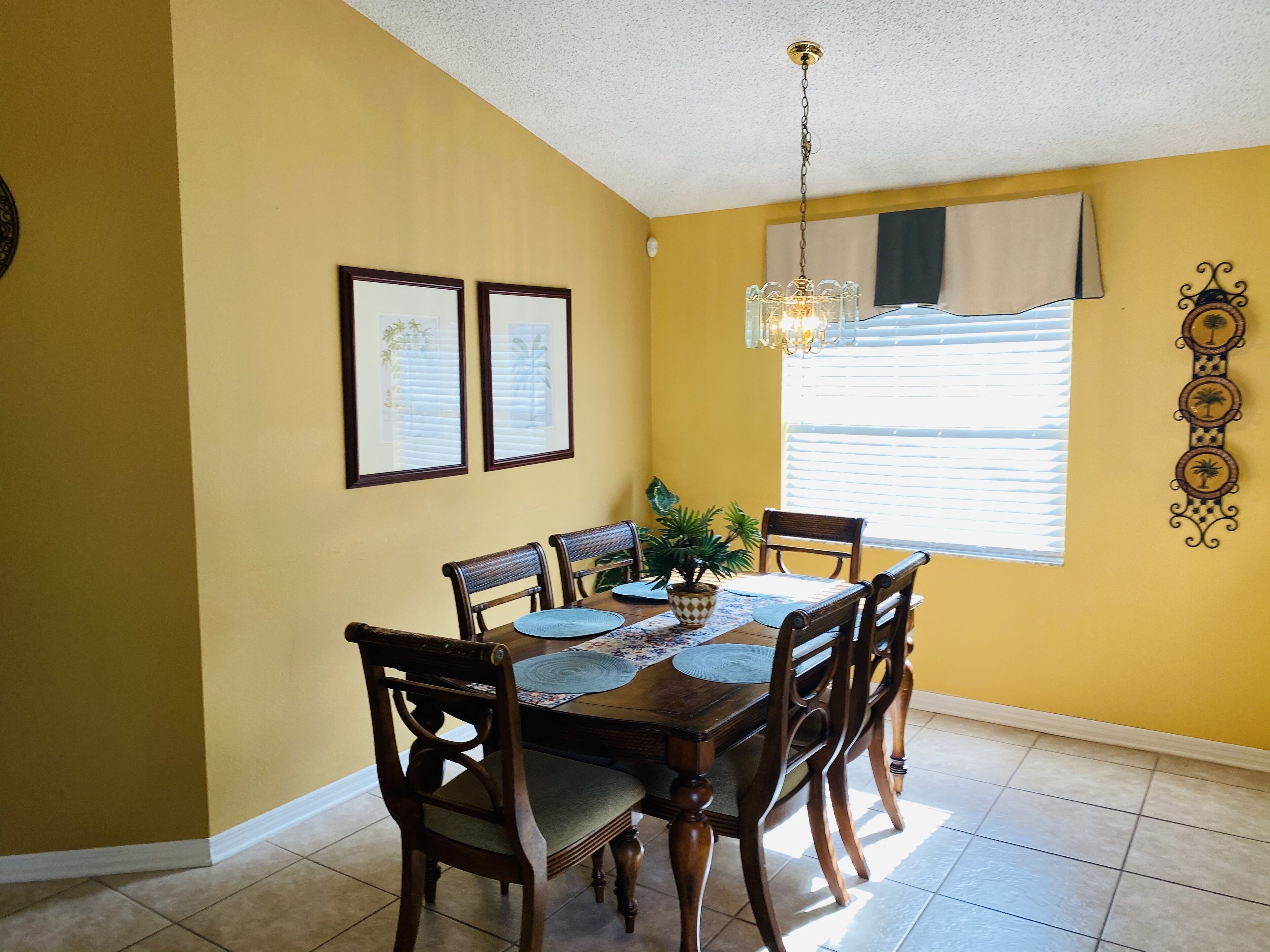 Un comedor con una mesa y sillas de madera, cuadros en la pared, una l ámpara colgante y una ventana con persianas.