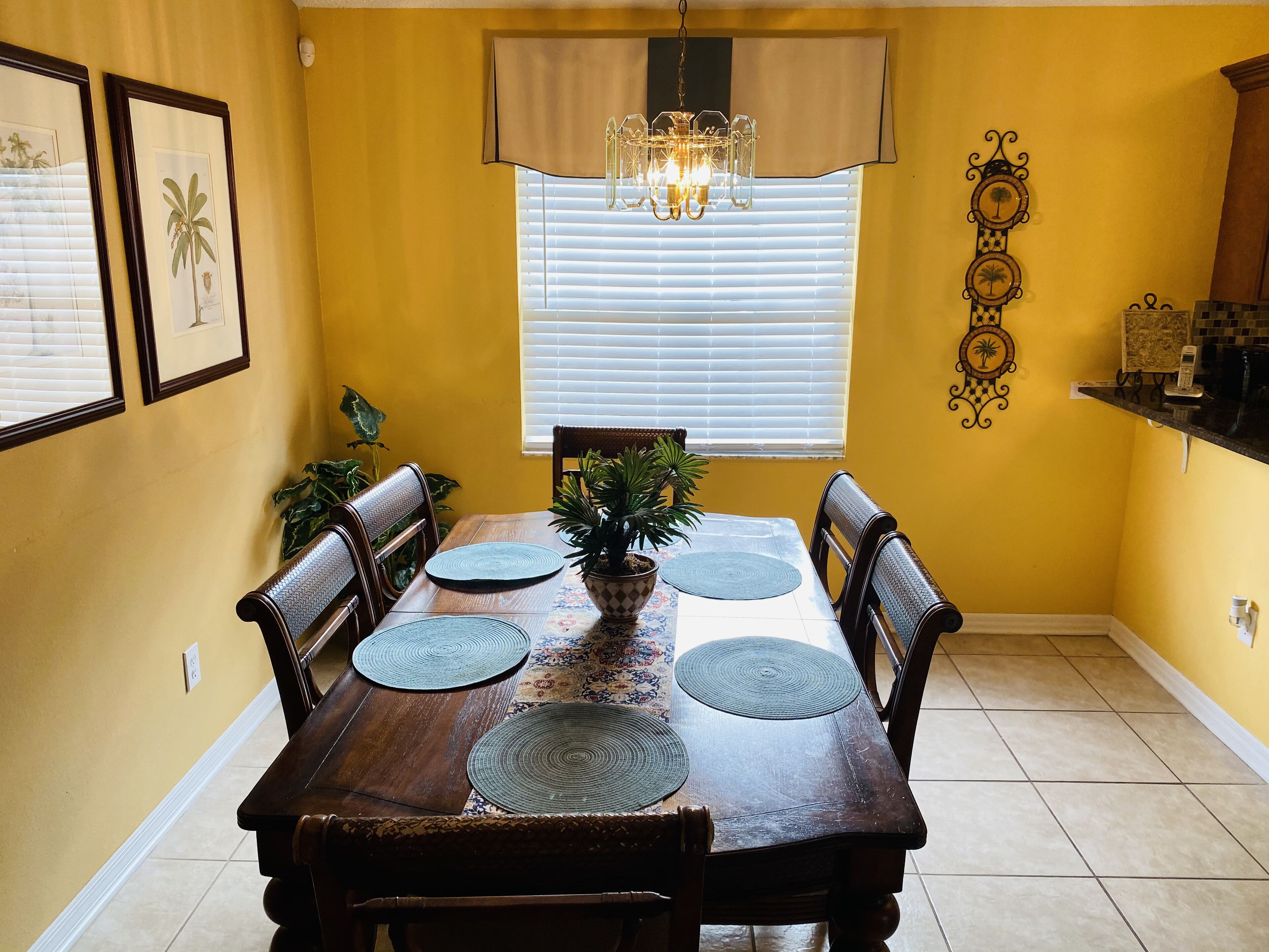 A dining room with a wooden table, six chairs, a chandelier, and a window with blinds.