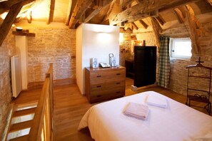 Ein gemütliches Schlafzimmer im Dachgeschoss mit freiliegenden Holzbalken, einer Steinwand, einer Holzkommode, einem Bett mit Handtüchern und einem Fenster mit Vorhängen.