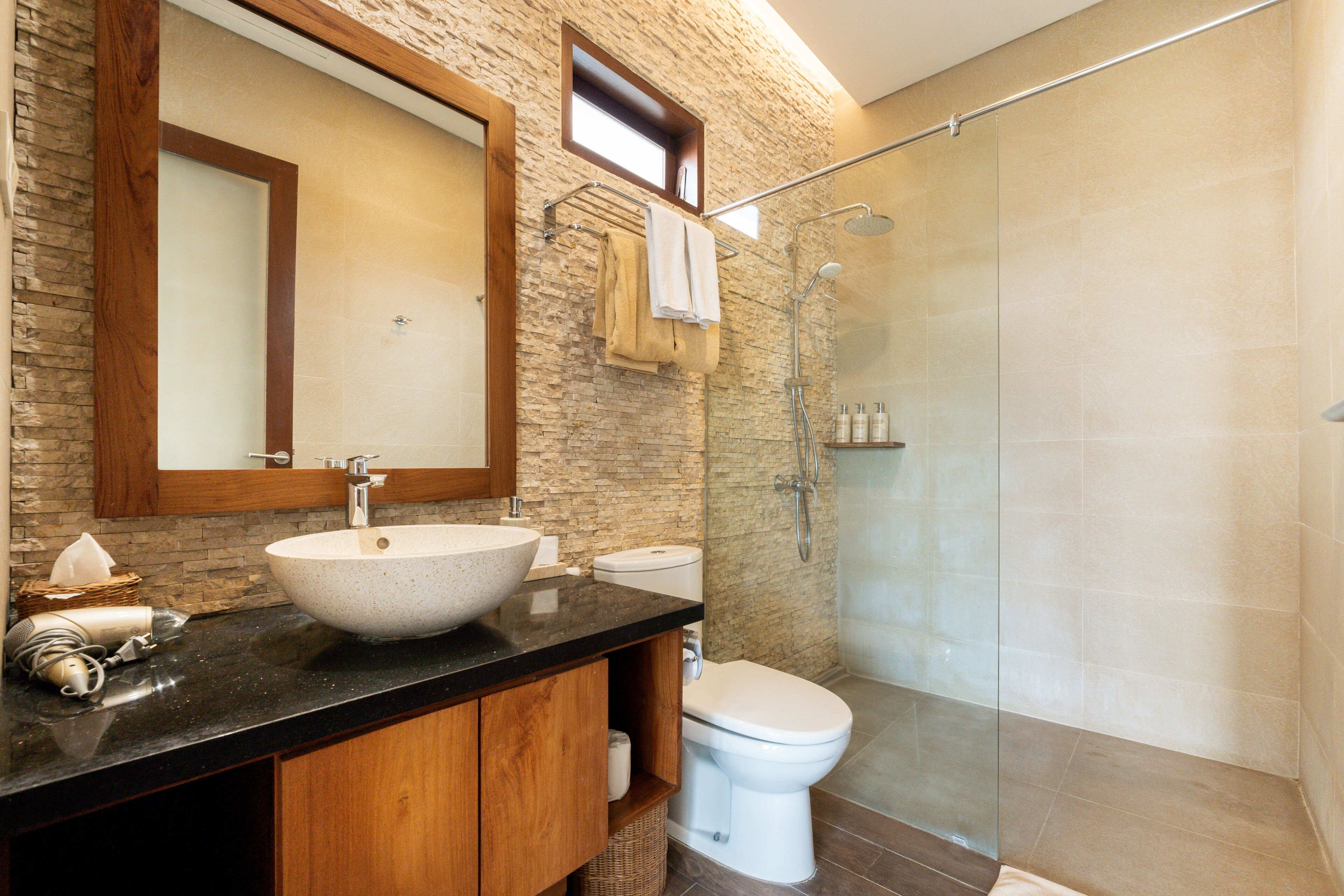 Un baño con pared de piedra, tocador de madera, un lavabo grande, inodoro y ducha con mampara de vidrio.