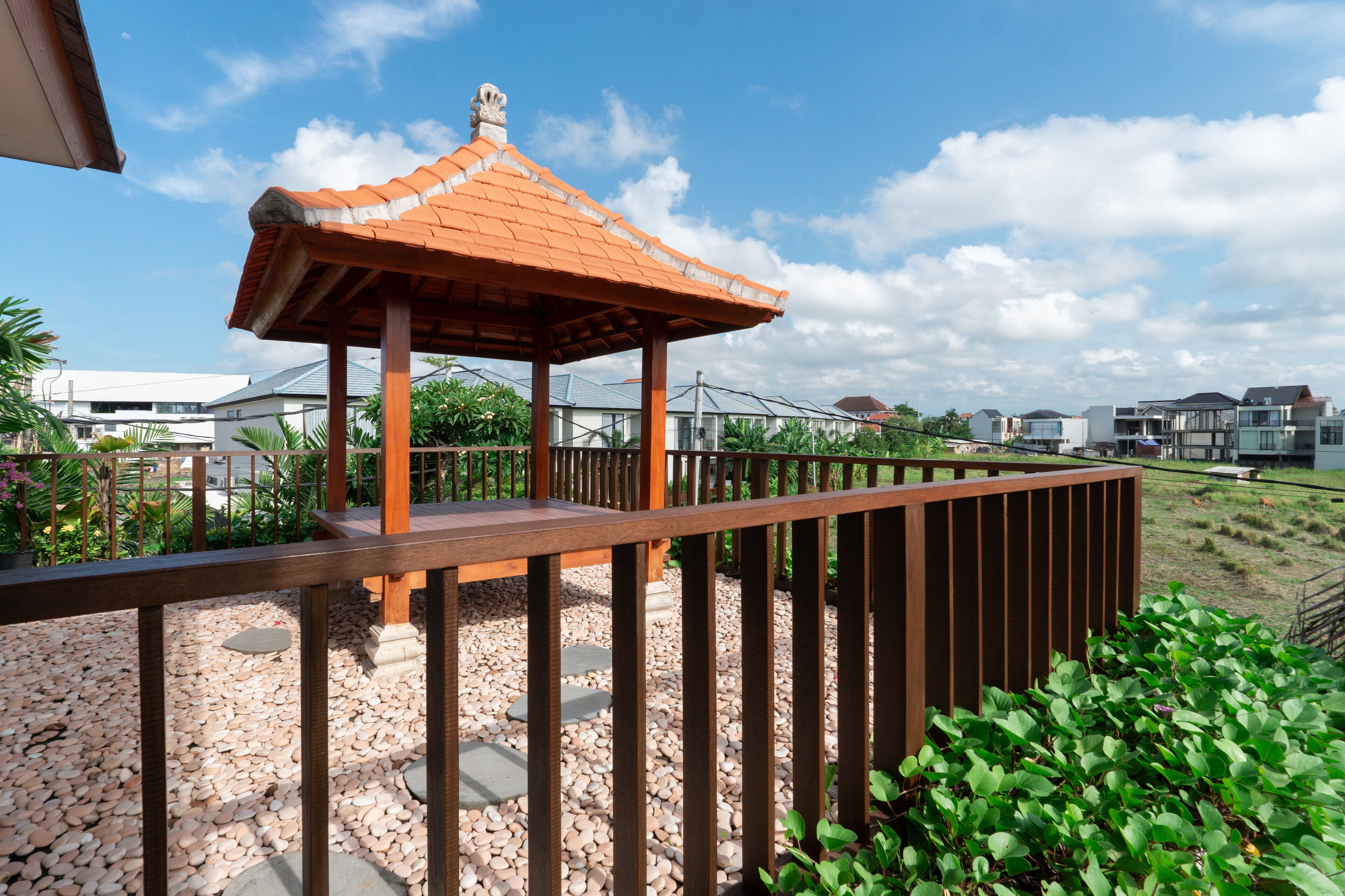 A gazebo with a tiled roof, surrounded by a wooden fence and pebbles, with modern buildings in the background.