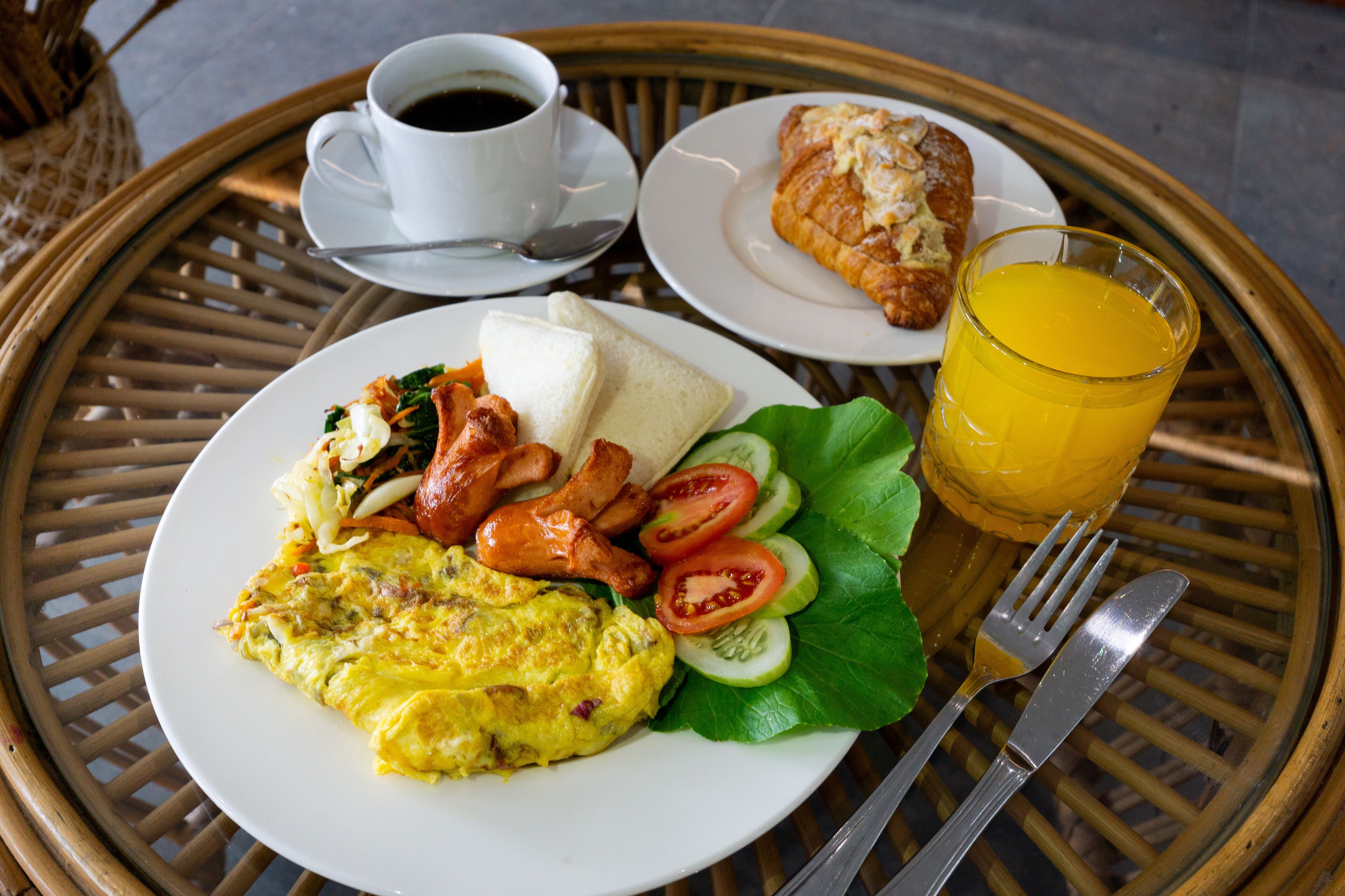 A breakfast setting with a plate of eggs, bacon, and a croissant, a cup of coffee, and a glass of orange juice on a wicker table.