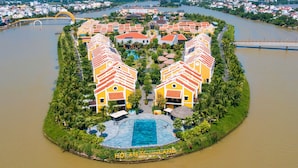 A resort island with multiple yellow buildings, a central swimming pool, and a river in the background. - Hoi An Memories Resort & Spa (Da Nang)
