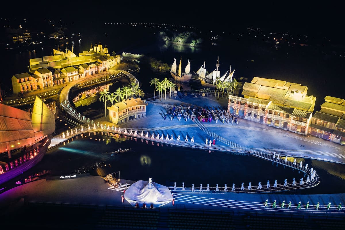 A nighttime view of a waterfront area with a large, illuminated building complex, a circular pedestrian bridge, and a central fountain.
